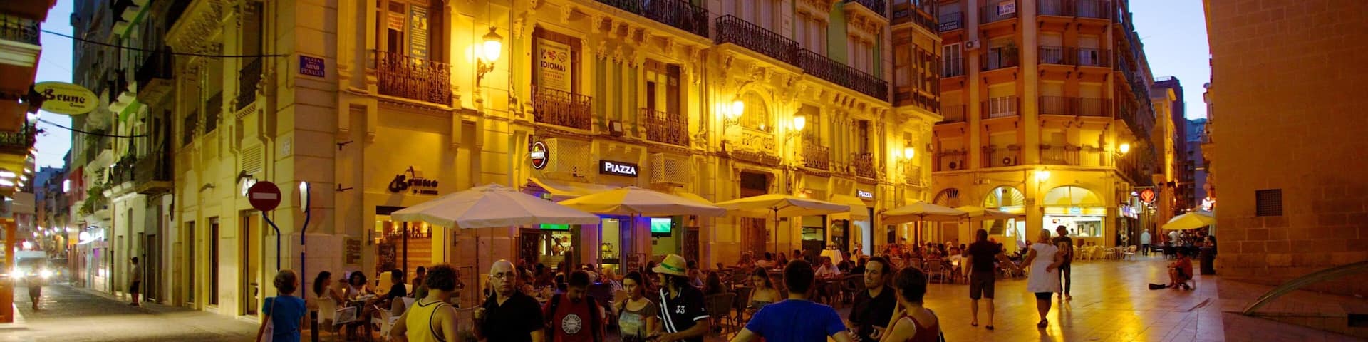 A bustling square with people walking and sitting at outdoor cafes, surrounded by historic buildings with ornate facades and balconies.