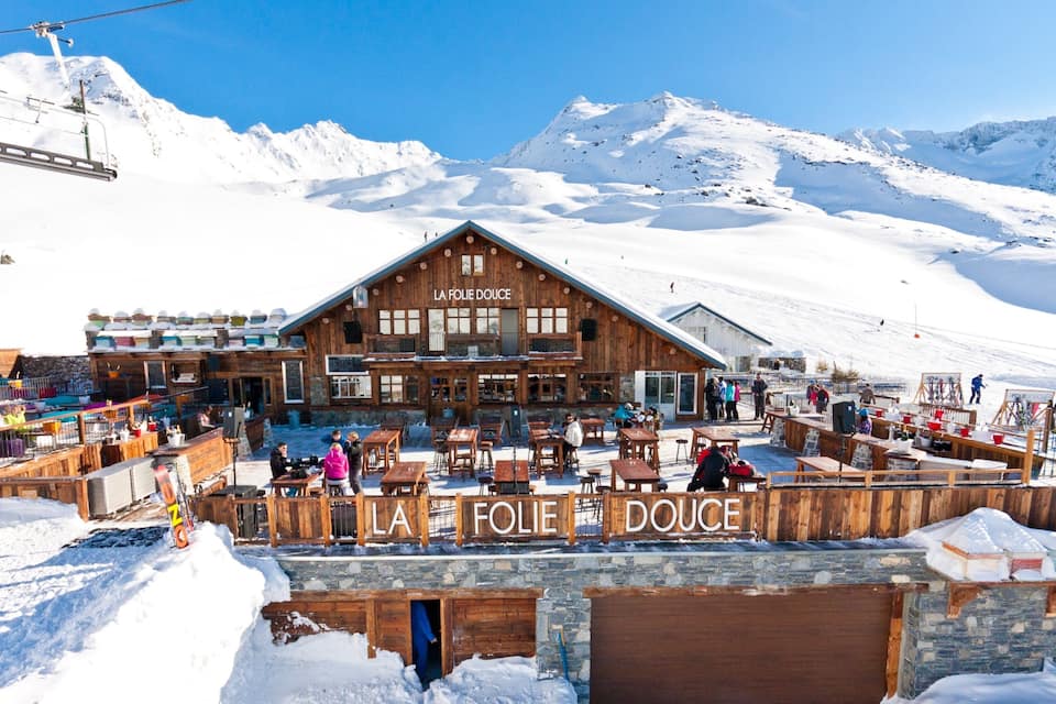 Un edificio in legno chiamato "LA FOLIE DOUCE" con posti a sedere all'aperto e montagne innevate sullo sfondo.