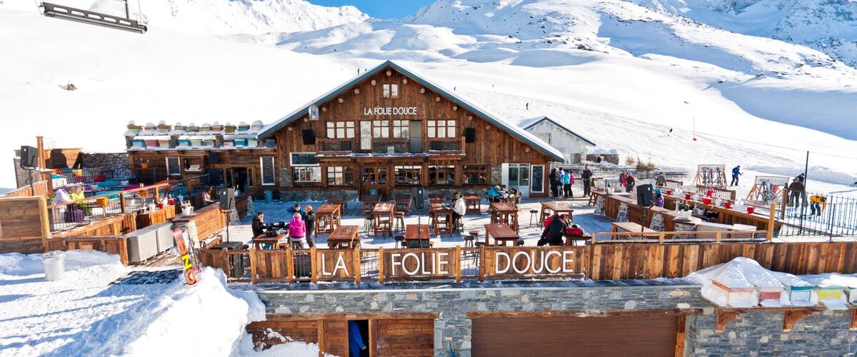 A wooden building named "LA FOLIE DOUCE" with outdoor seating and snow-covered mountains in the background.