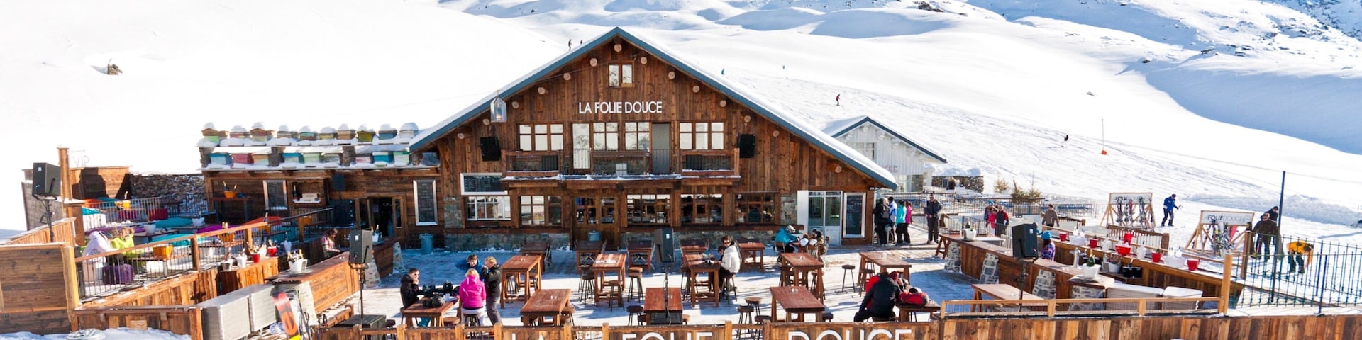 A wooden building named "LA FOLIE DOUCE" with outdoor seating and snow-covered mountains in the background.