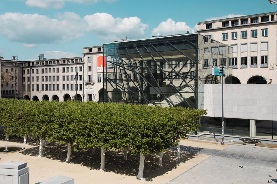 A modern building with a glass facade and metal framework, surrounded by a row of trees and a paved area.
