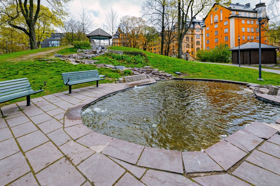 A park with a pond, benches, a gazebo, and buildings in the background.