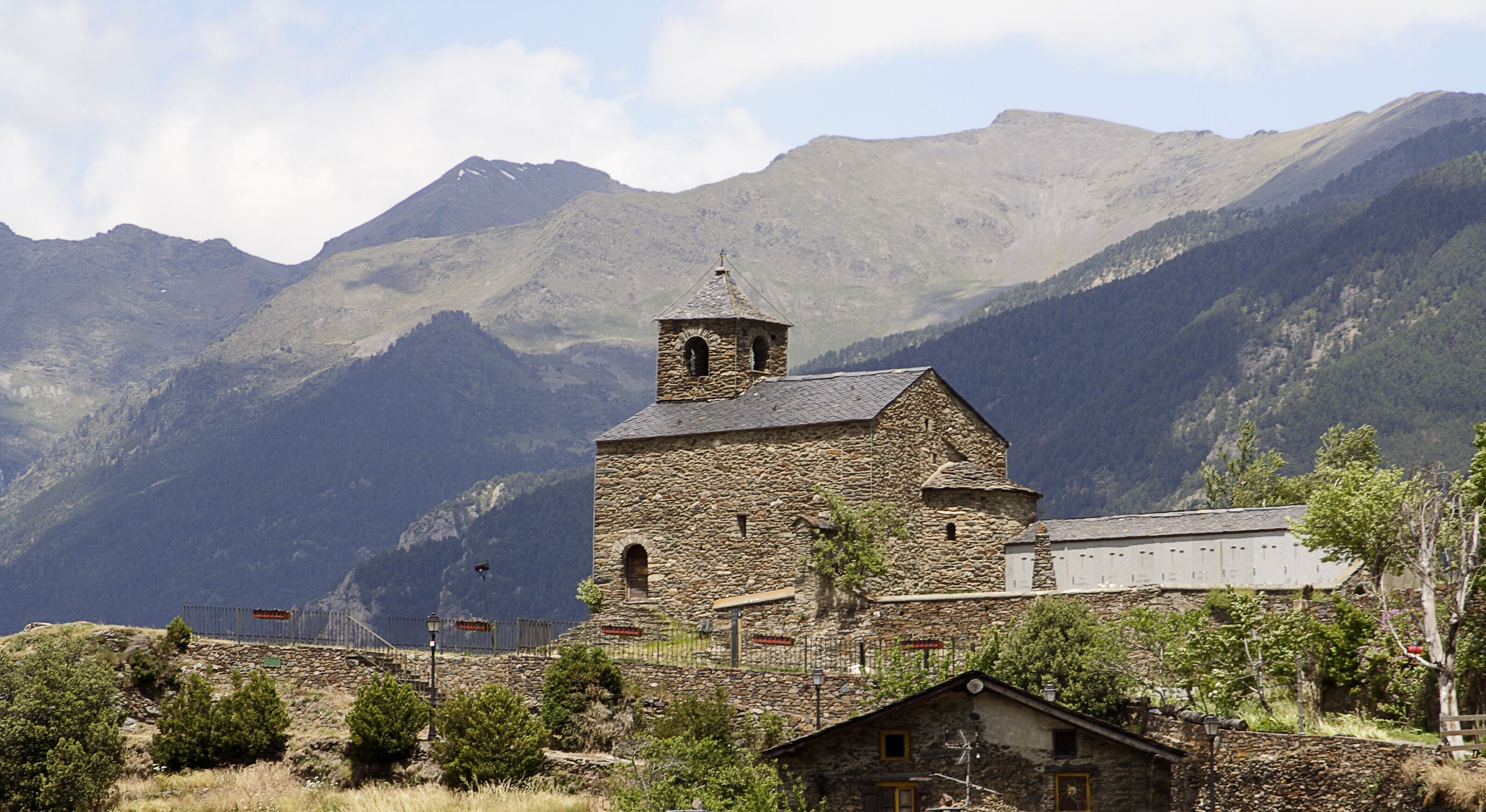 A stone church with a bell tower, surrounded by mountains and a small village.