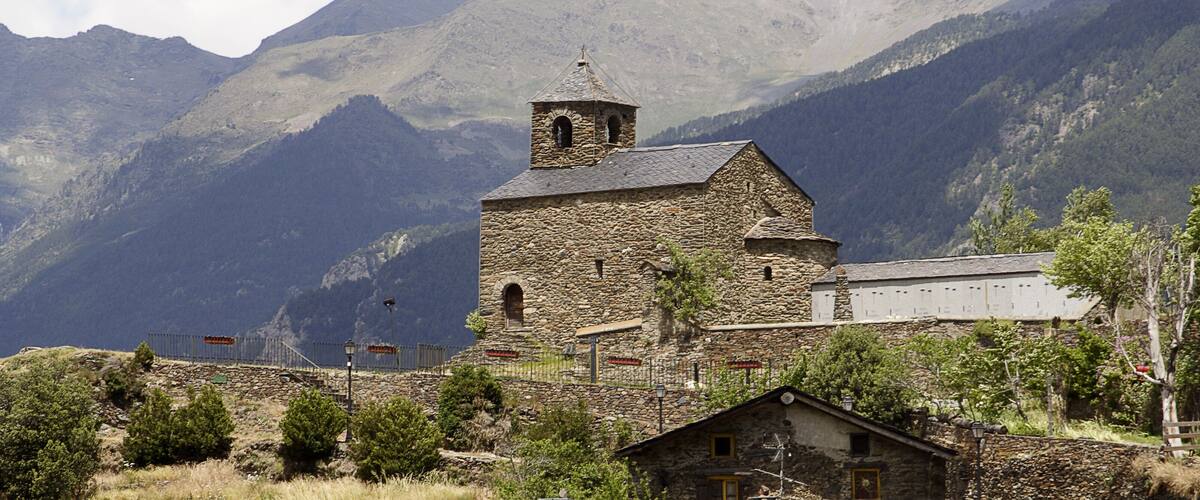 A stone church with a bell tower, surrounded by mountains and a small village.