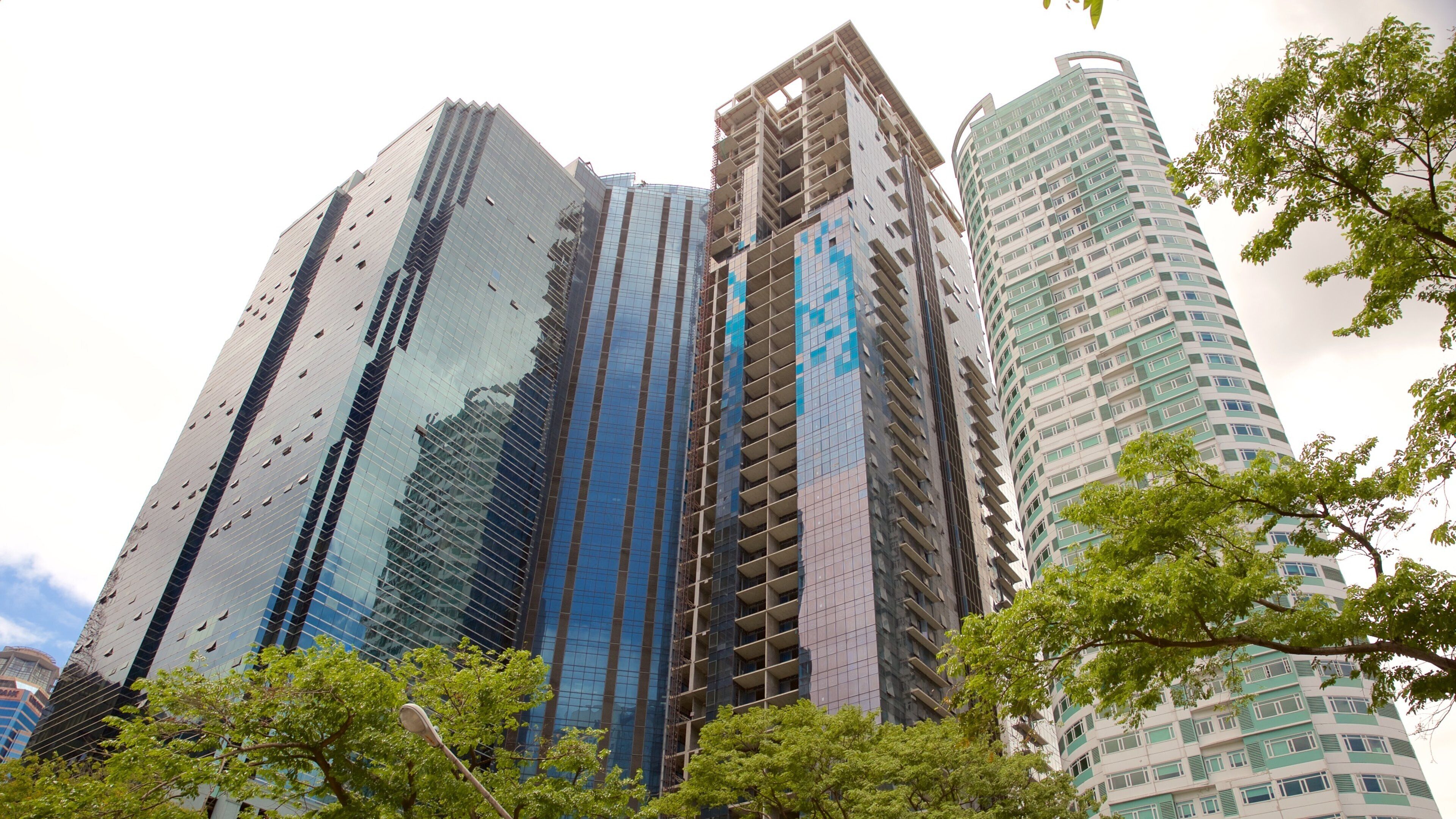 A cluster of high-rise buildings with reflective glass facades, some under construction, and surrounded by green trees.