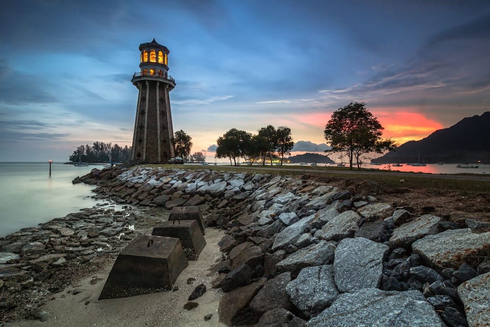 Un faro, costa rocosa, árboles y un atardecer sobre el agua.