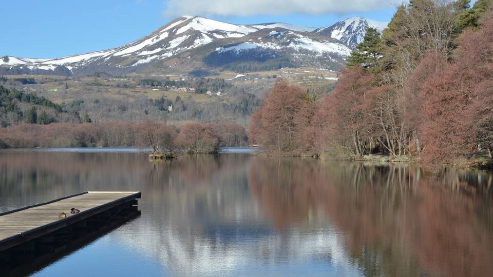 A calm lake with a wooden dock, surrounded by autumn-colored trees and a snow-capped mountain in the background.