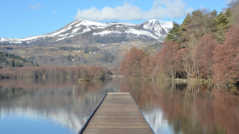 A wooden dock extending into a calm lake with a backdrop of snow-capped mountains and autumn-colored trees.