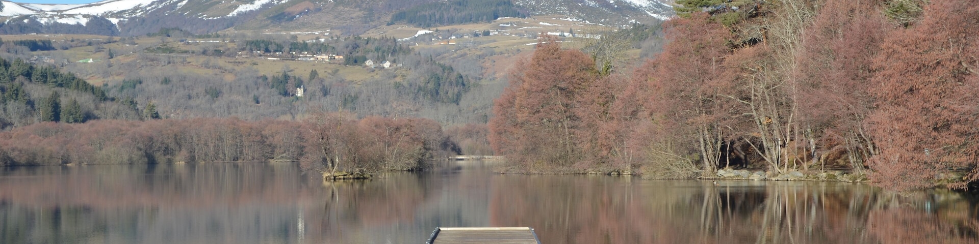 Un muelle de madera se adentra en un lago tranquilo, con montañas nevadas y árboles de colores otoñales al fondo.