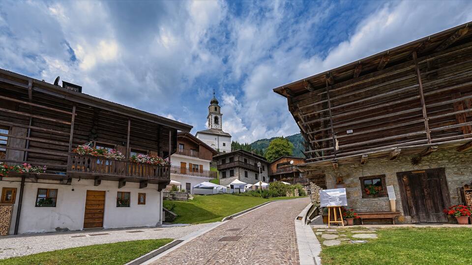 A cobblestone street in a village with wooden buildings, a church with a bell tower, and a mountain in the background.