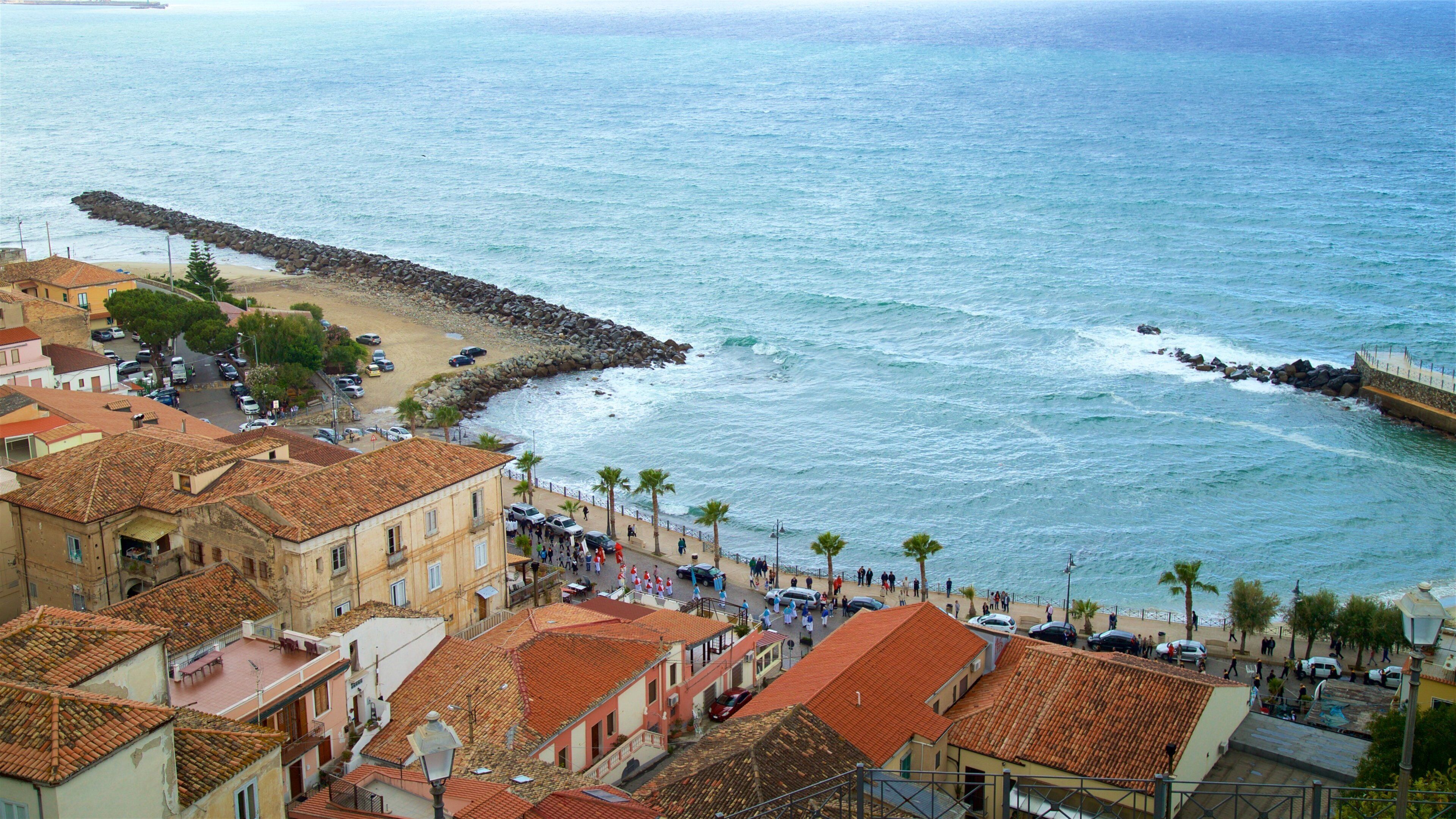 Uma cidade costeira com edifícios de telhados em terracota, um molhe rochoso e uma praia arenosa.