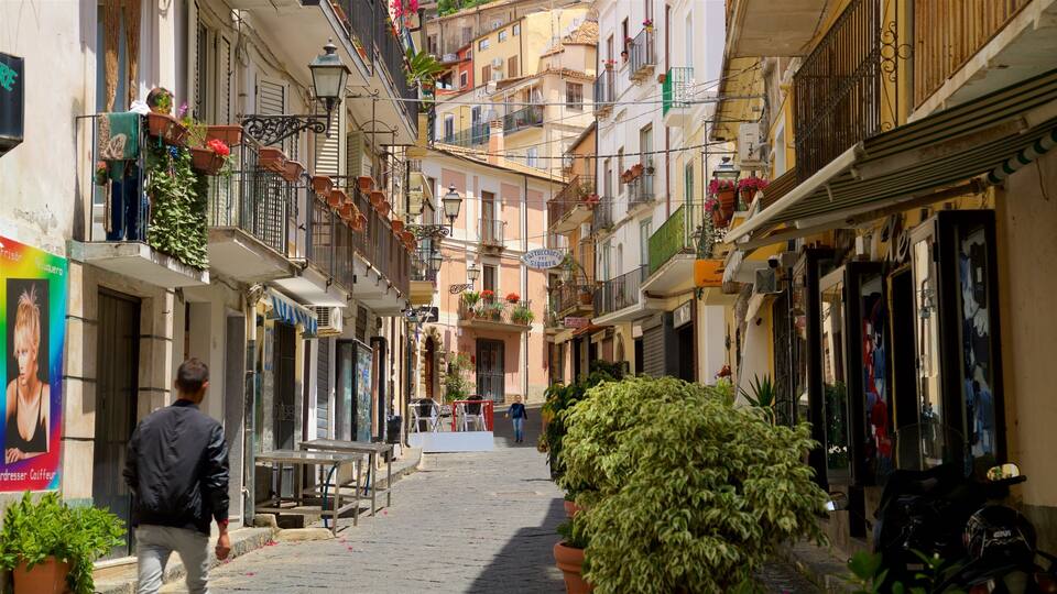 A narrow street with buildings on either side, balconies with plants, and a few pedestrians.