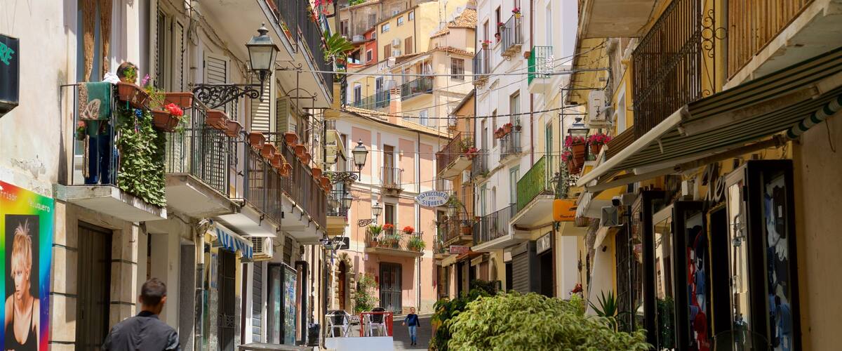 A narrow street with buildings on either side, balconies with plants, and a few pedestrians.