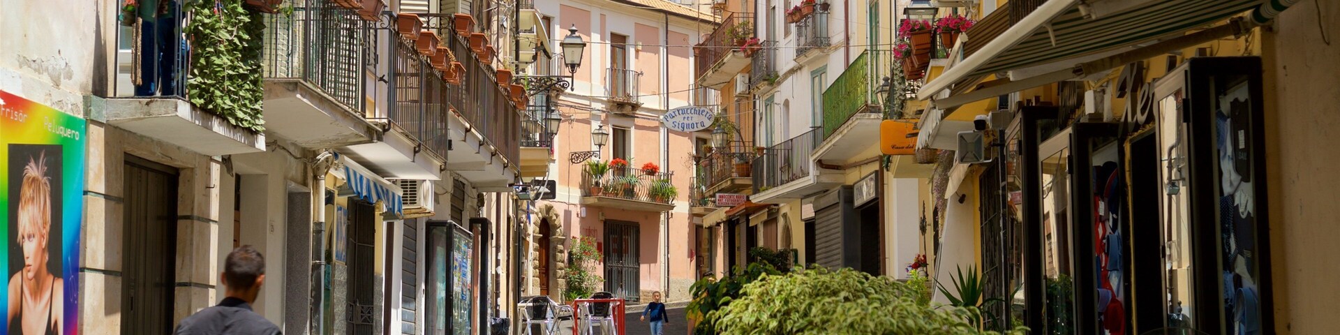 A narrow street with buildings on either side, balconies with plants, and a few pedestrians.