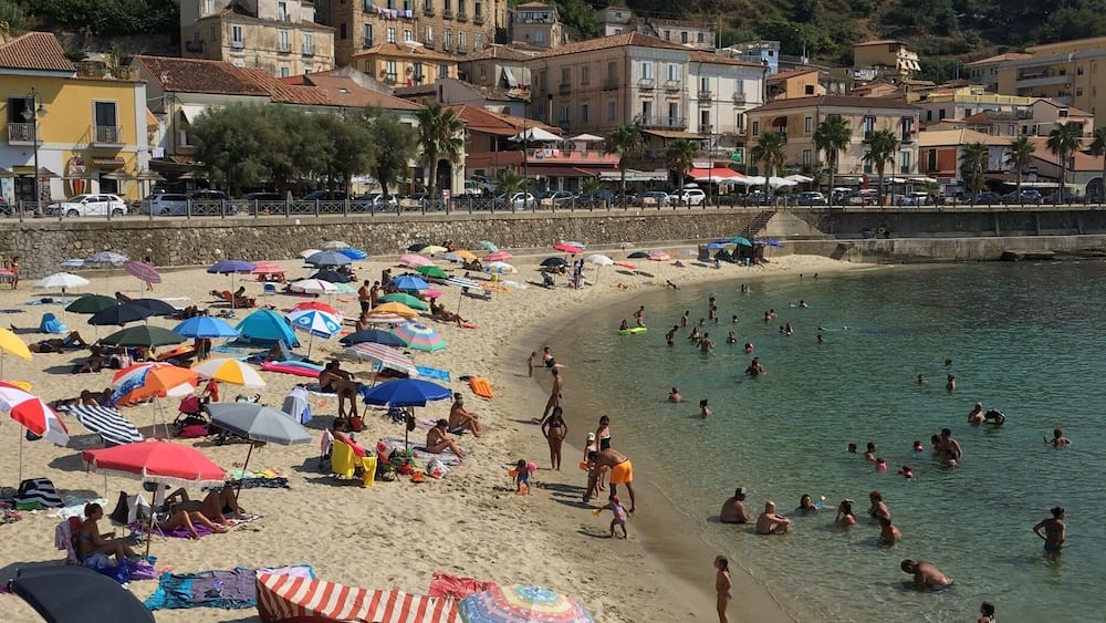 A crowded beach with people swimming and sunbathing, surrounded by colorful umbrellas and beach chairs.