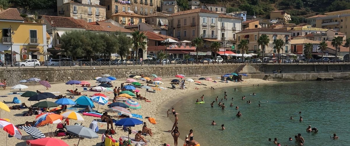 A crowded beach with people swimming and sunbathing, surrounded by colorful umbrellas and beach chairs.