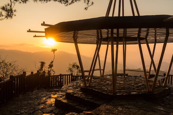 A wooden structure with a metal roof, set against a sunset with mountains in the background.