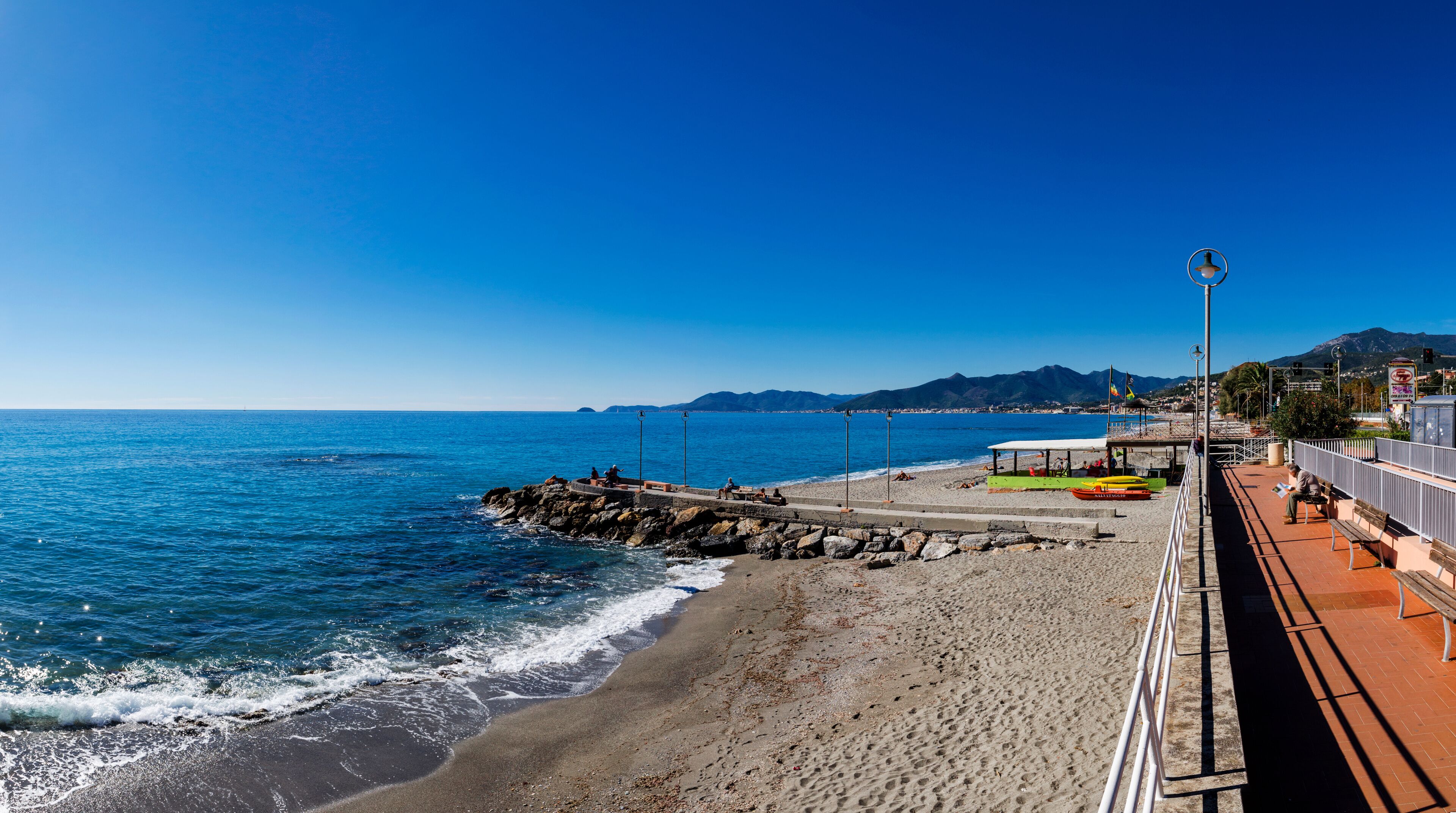 A coastal promenade with a rocky breakwater, beach, and a clear blue sky.