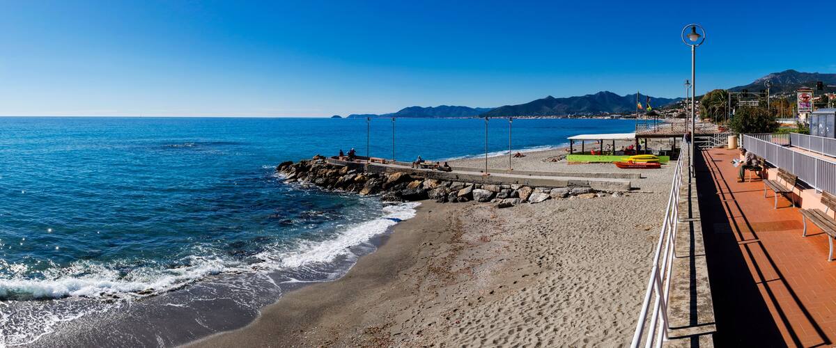 A coastal promenade with a rocky breakwater, beach, and a clear blue sky.
