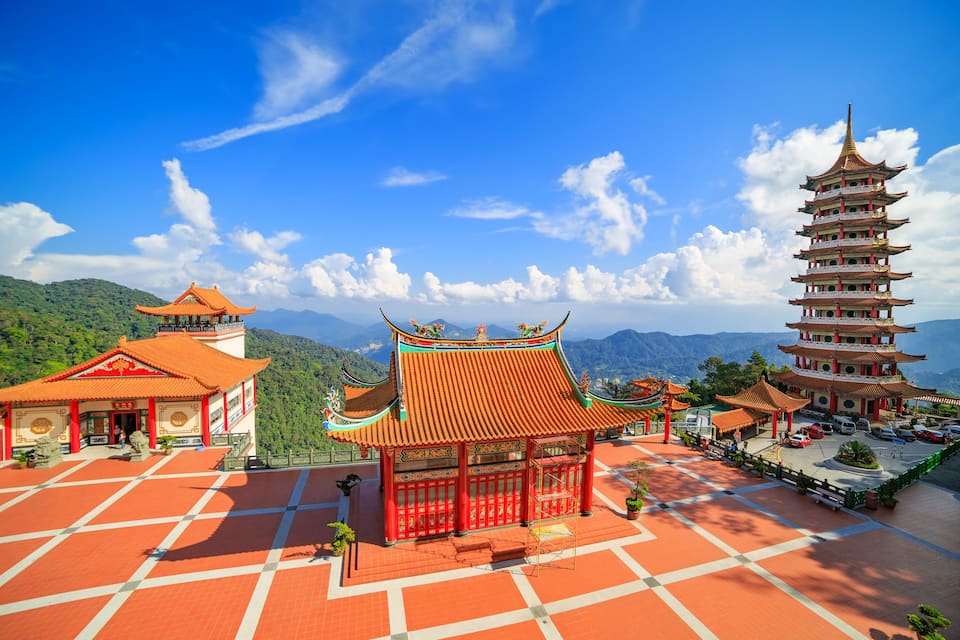 A traditional Chinese pagoda and pavilion complex with terracotta roofs and red accents, set against a backdrop of mountains and a clear blue sky.