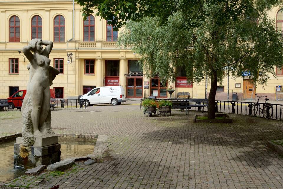 Una estatua, una fuente, una camioneta blanca, bancos y un edificio con ventanas arqueadas.