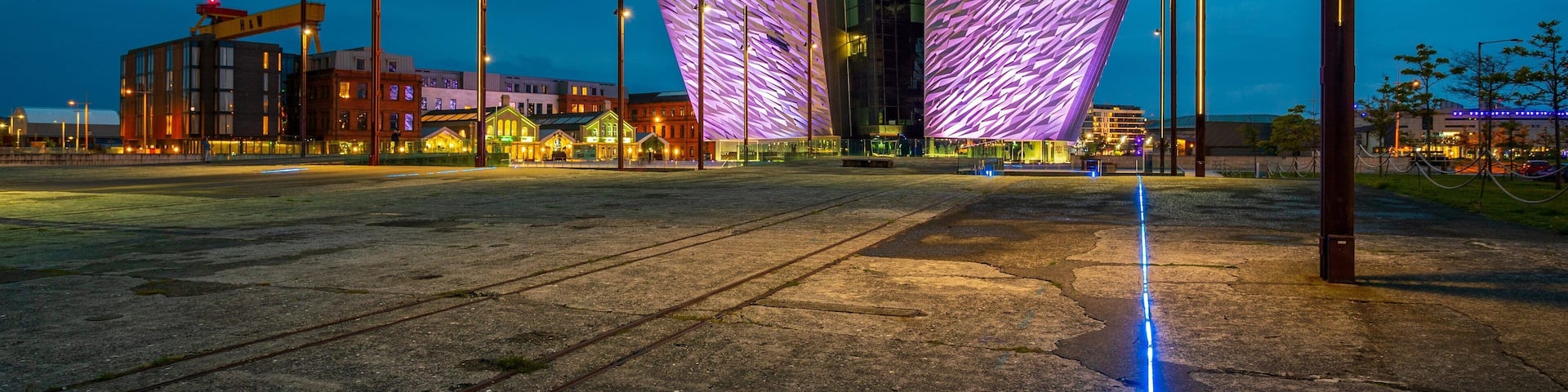A modern building with a unique purple-lit facade and a large open plaza with a train track.