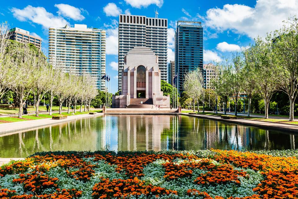 Un parc amb un estany que reflecteix, llits de flors de colors i un monument central envoltat d'edificis alts.
