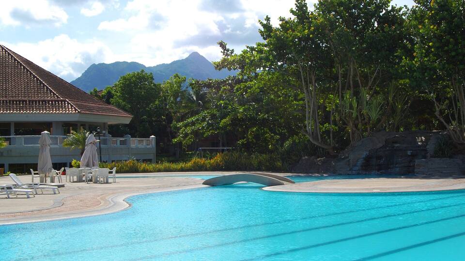 A swimming pool with lounge chairs and umbrellas, surrounded by trees and a building with a tiled roof.