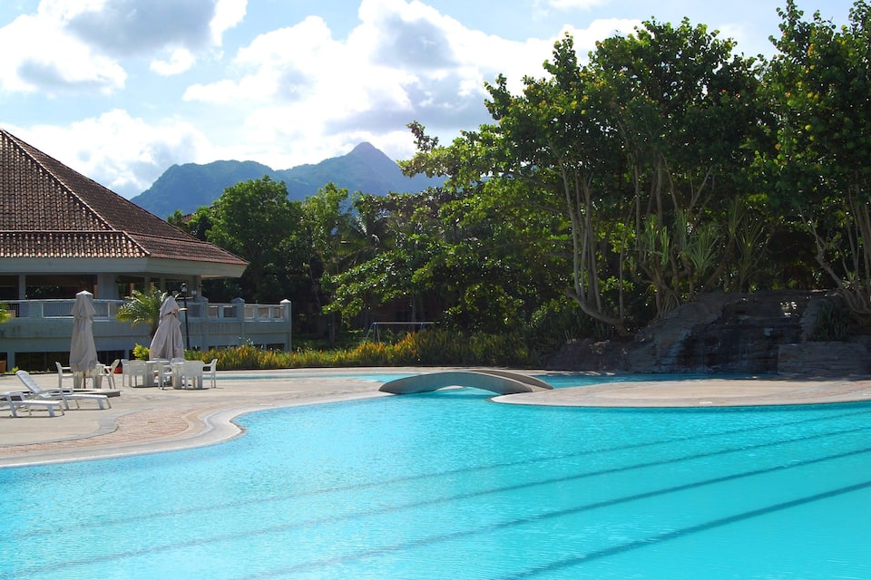 A swimming pool with lounge chairs and umbrellas, surrounded by trees and a building with a tiled roof.