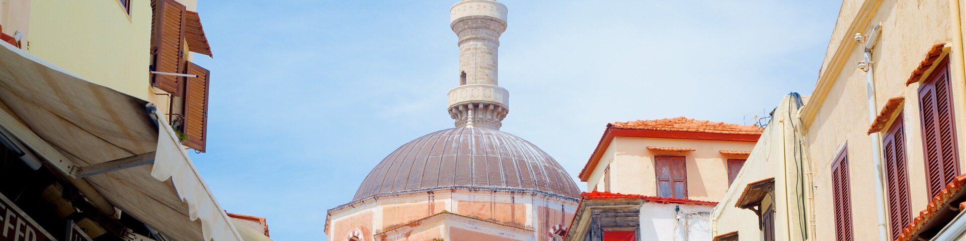 Uma mesquita com um minarete alto e uma cúpula, cercada por ruas estreitas com lojas.