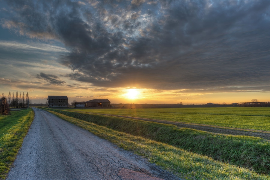 Een landelijk landschap met een onverharde weg, velden en schuren onder een bewolkte lucht bij zonsondergang.