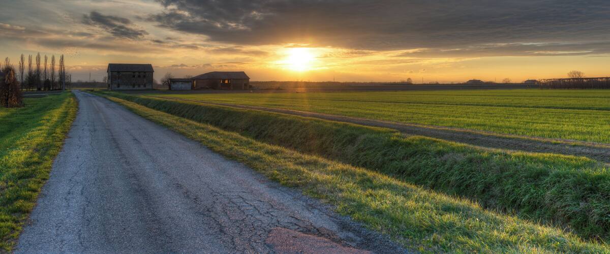 A rural landscape with a dirt road, fields, and barns under a cloudy sky at sunset.