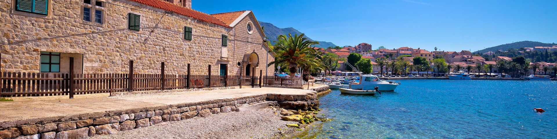 Un edificio de piedra con techo de tejas rojas, un frente marítimo despejado y barcos amarrados cerca de la orilla.