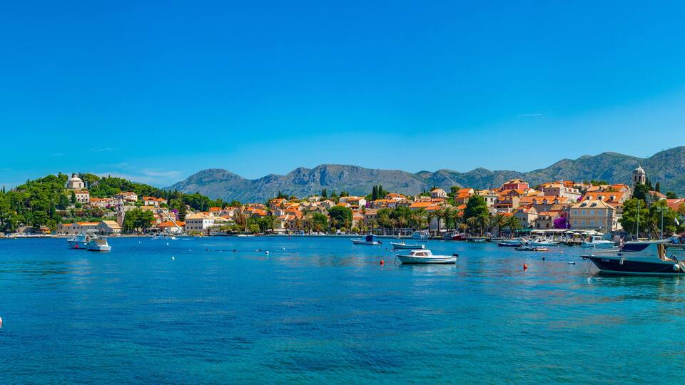 A coastal town with boats, a clear blue sea, and mountains in the background.