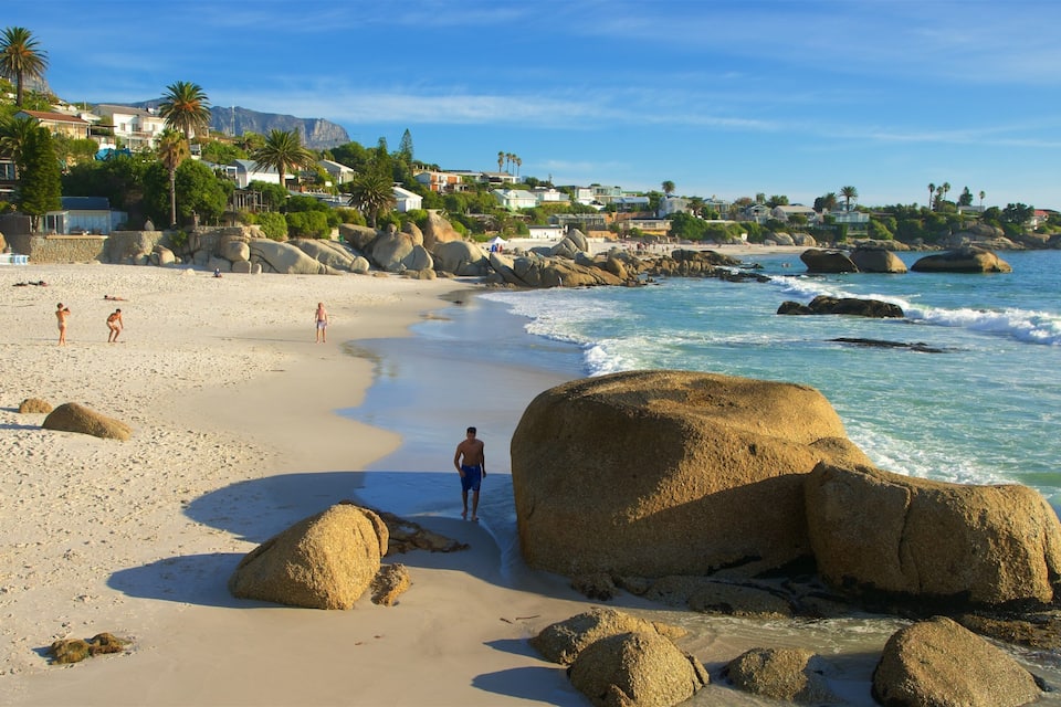 A sandy beach with large rocks, calm waves, and houses in the background.
