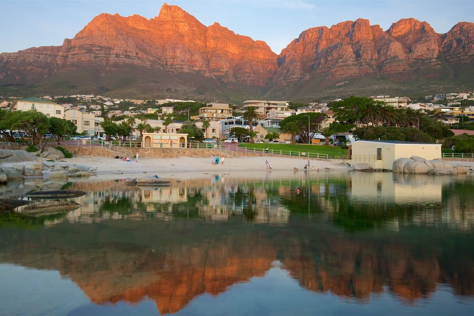 A coastal town with a rocky mountain range in the background, calm water reflecting the scenery, and a sandy beach with people and buildings.