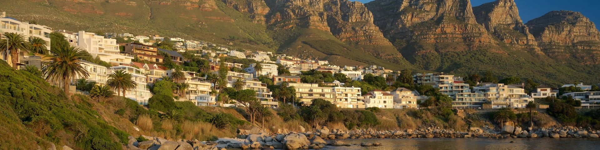 A coastal town with buildings, rocky shoreline, and mountains in the background.