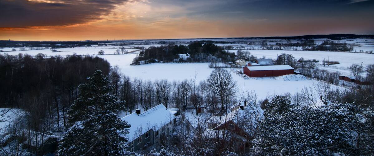 A snowy landscape with a red barn, trees, and a sunset sky.