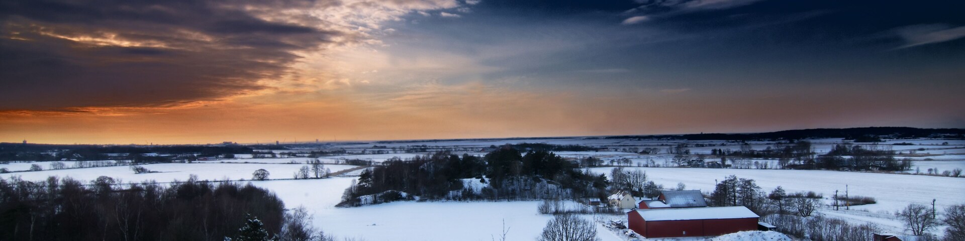 A snowy landscape with a red barn, trees, and a sunset sky.