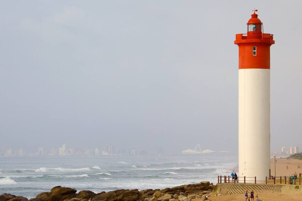 Ein hoher, rot-weiß gestreifter Leuchtturm an der Küste, wo Wellen gegen Felsen schlagen.
