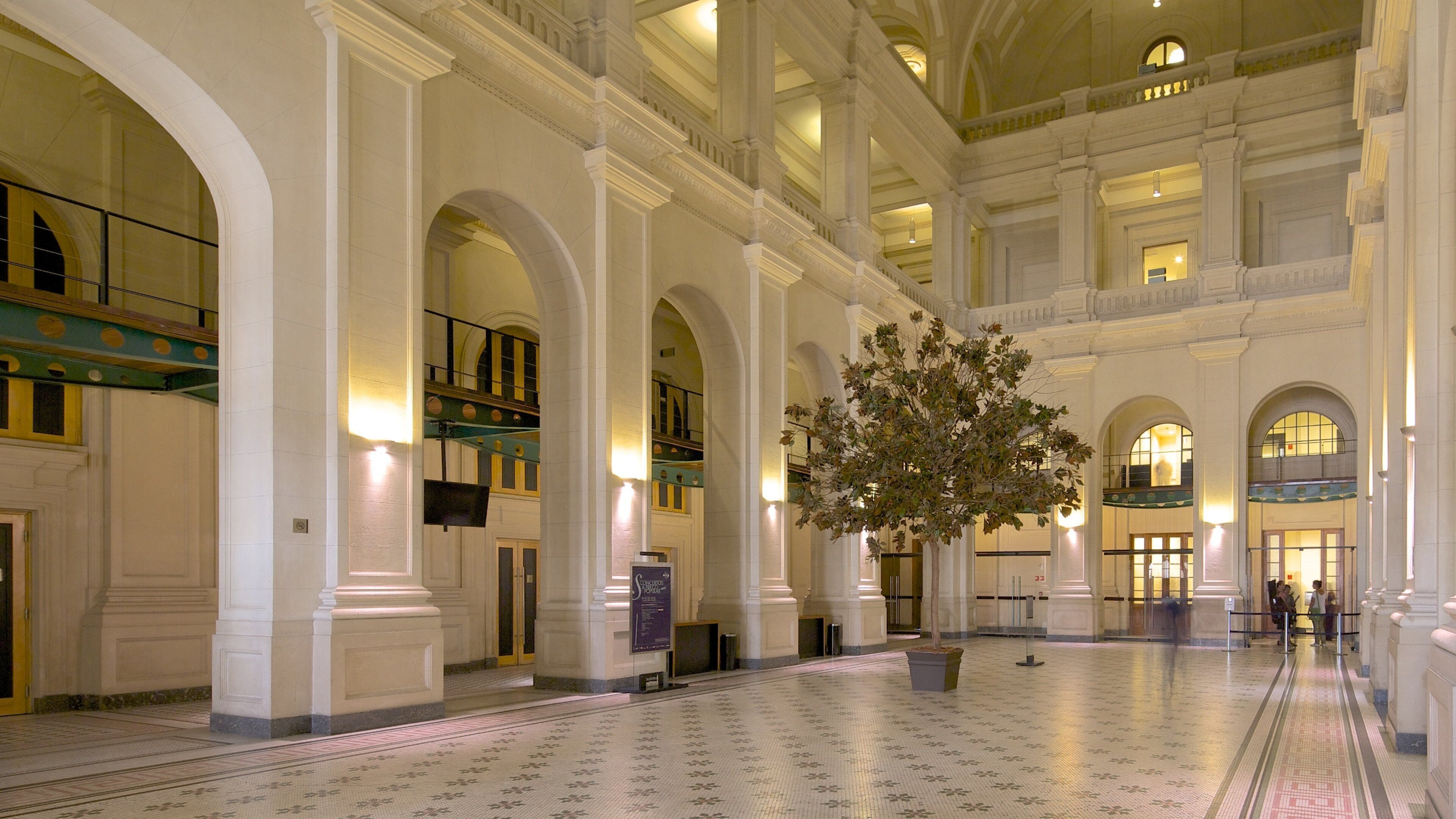 A grand hall with high ceilings, arched doorways, and a large tree in the center.