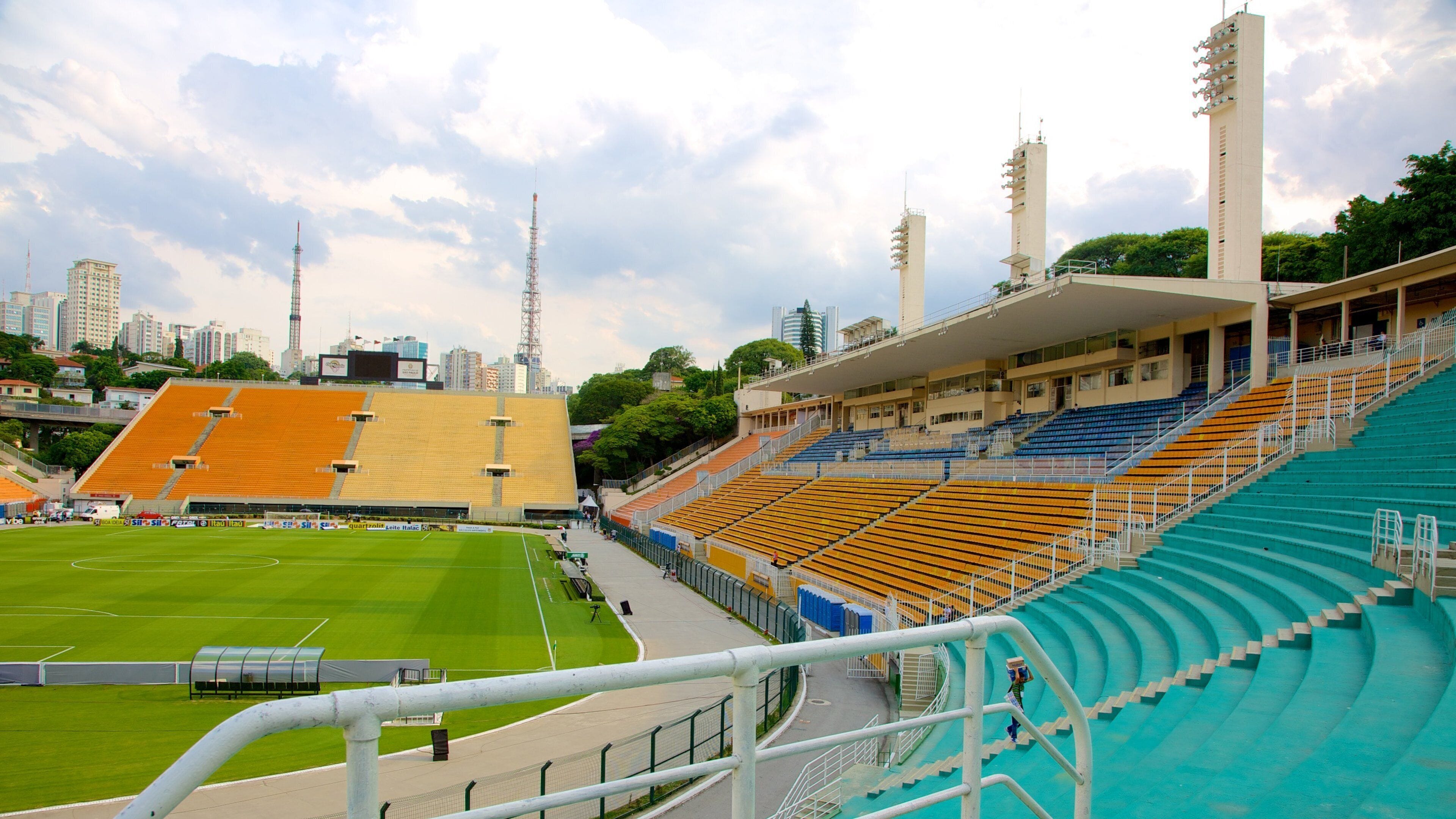 Um estádio com arquibancadas em níveis, um campo de futebol e um horizonte urbano ao fundo.