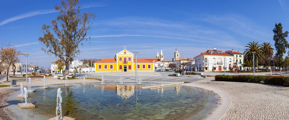 A plaza with a fountain, yellow buildings, and a church with a red roof.