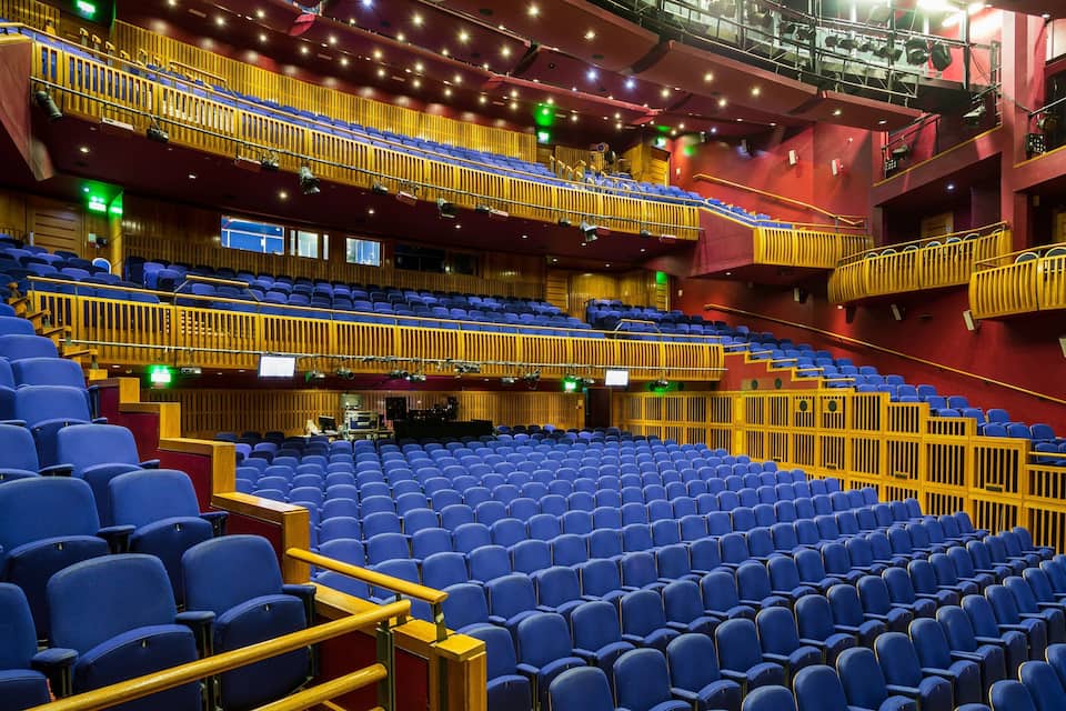 A large indoor auditorium with blue seats, wooden railings, and a stage area.