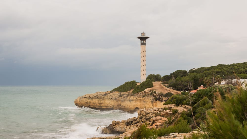 A coastal landscape with a lighthouse on a rocky outcrop, surrounded by greenery and a clear sky.