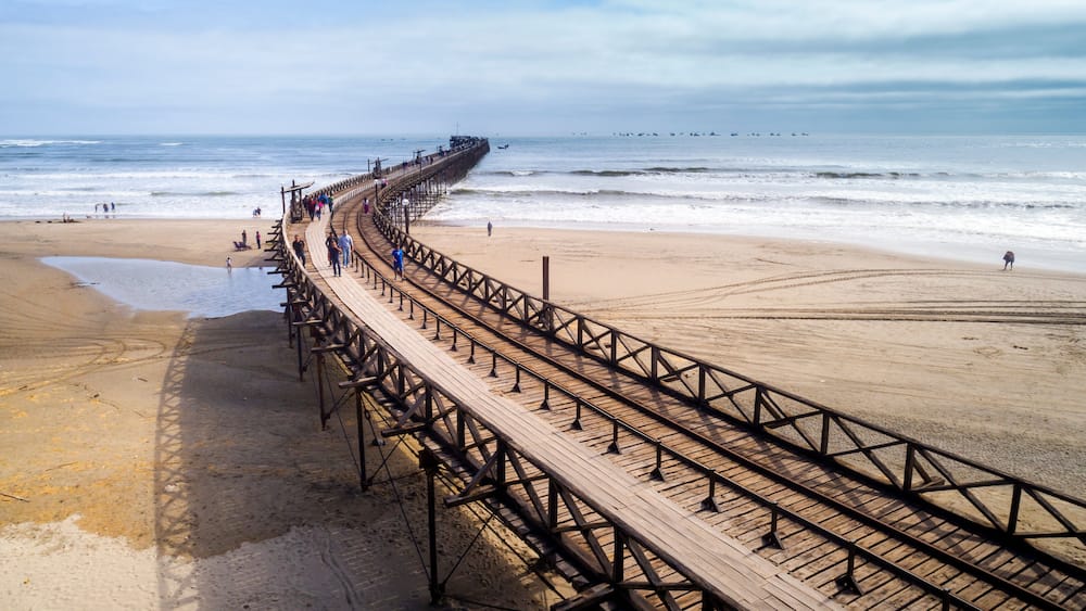 Eine hölzerne Promenade erstreckt sich ins Meer, Menschen laufen darauf.