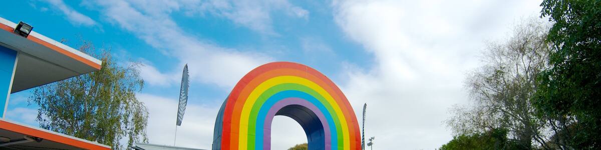 Un arc-en-ciel coloré en forme de porte d’entrée orne l’entrée d’un parc, avec un panneau indicateur et des personnes debout à proximité.