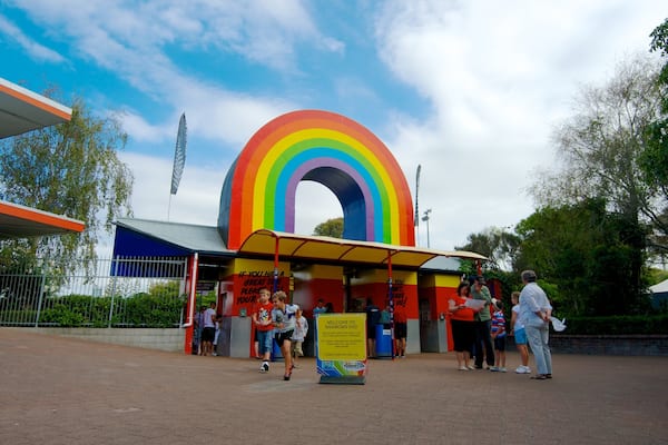 Un arc-en-ciel coloré en forme de porte d’entrée orne l’entrée d’un parc, avec un panneau indicateur et des personnes debout à proximité.