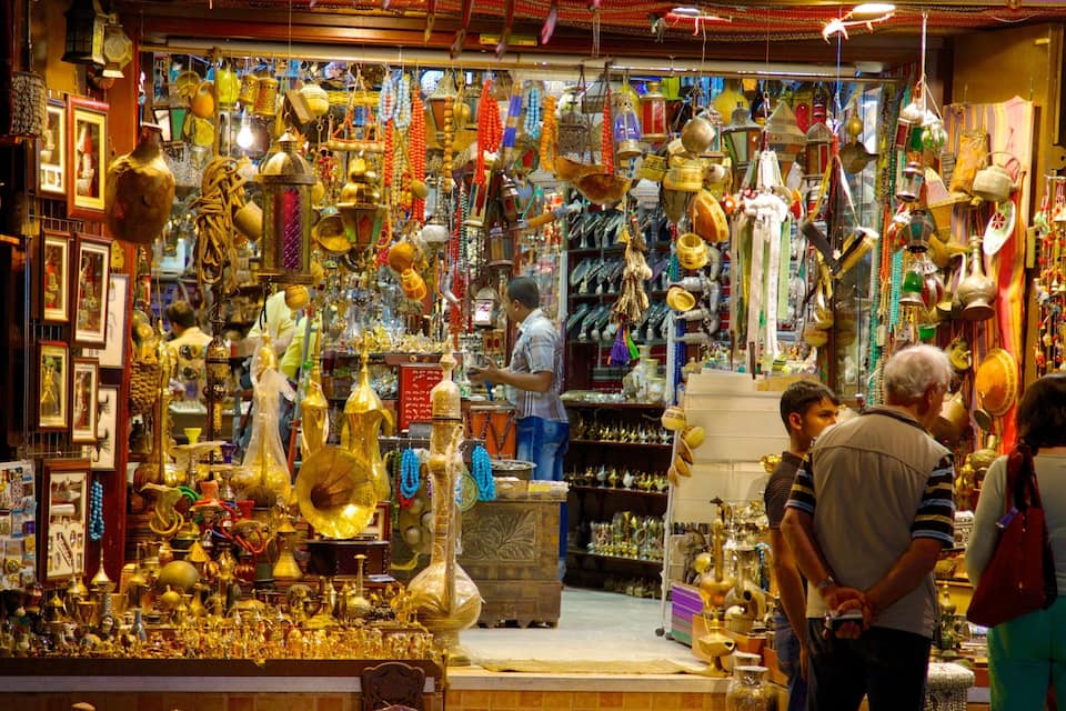 Uma banca de mercado movimentada repleta de diversos objetos de ouro e prata, incluindo vasos, tigelas e joias.