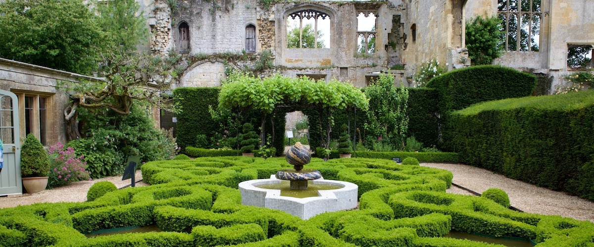 A formal garden with a central fountain, trimmed hedges, and an old stone building in the background.